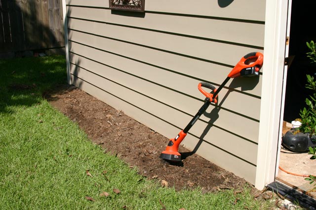 Black and Decker orange cordless tiller green grass garage vinyl siding and empty flower bed
