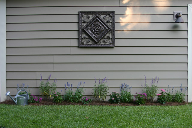 blue and purple salvia in flower bed against tan vinyl siding with brown medallion with pink and purl pentas