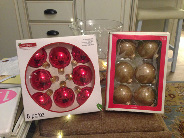 boxes of red and gold Christmas ornaments sitting on tan ottoman in sunroom